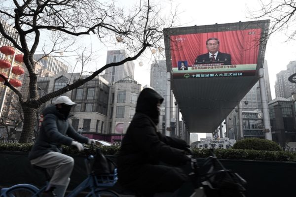 2025年3月5日，北京一家購物中心外，人們騎行經過現場直播總理李強在全國人大會議開幕式上講話的屏幕。(Greg Baker/AFP via Getty Images)
