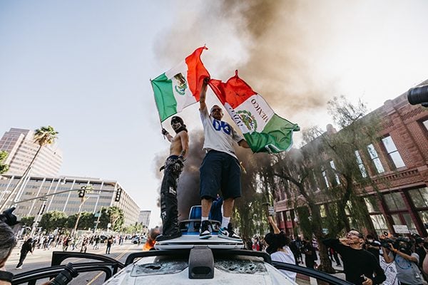 2025年6月8日，在加州洛杉矶爆发抗议期间，两名抗议者站在一辆被砸坏的Waymo汽车上，挥舞着墨西哥国旗。(David Pashaee/Middle East Images/AFP via Getty Images)
